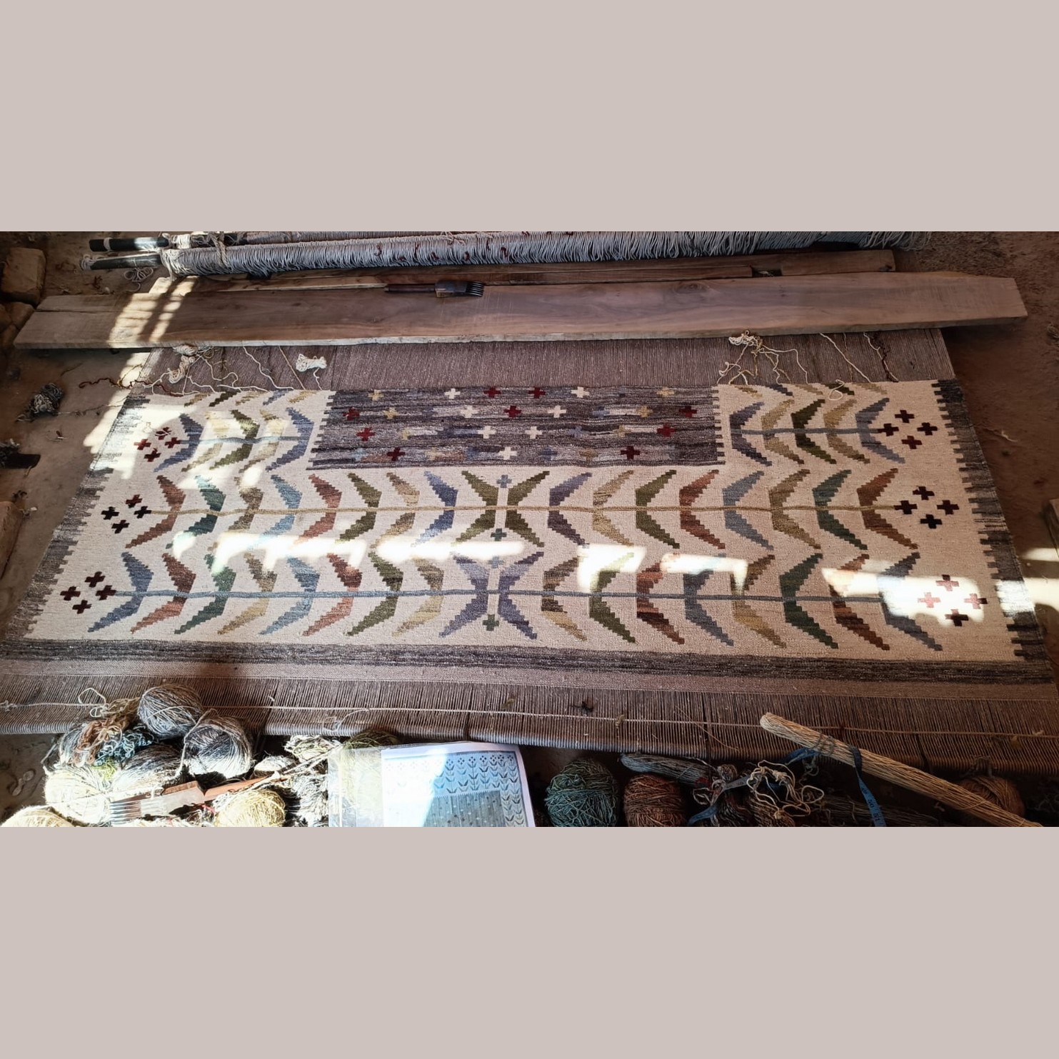 Grote geweven kelim met geometrische patronen, in een houten frame, met wol en tapijten eromheen.
