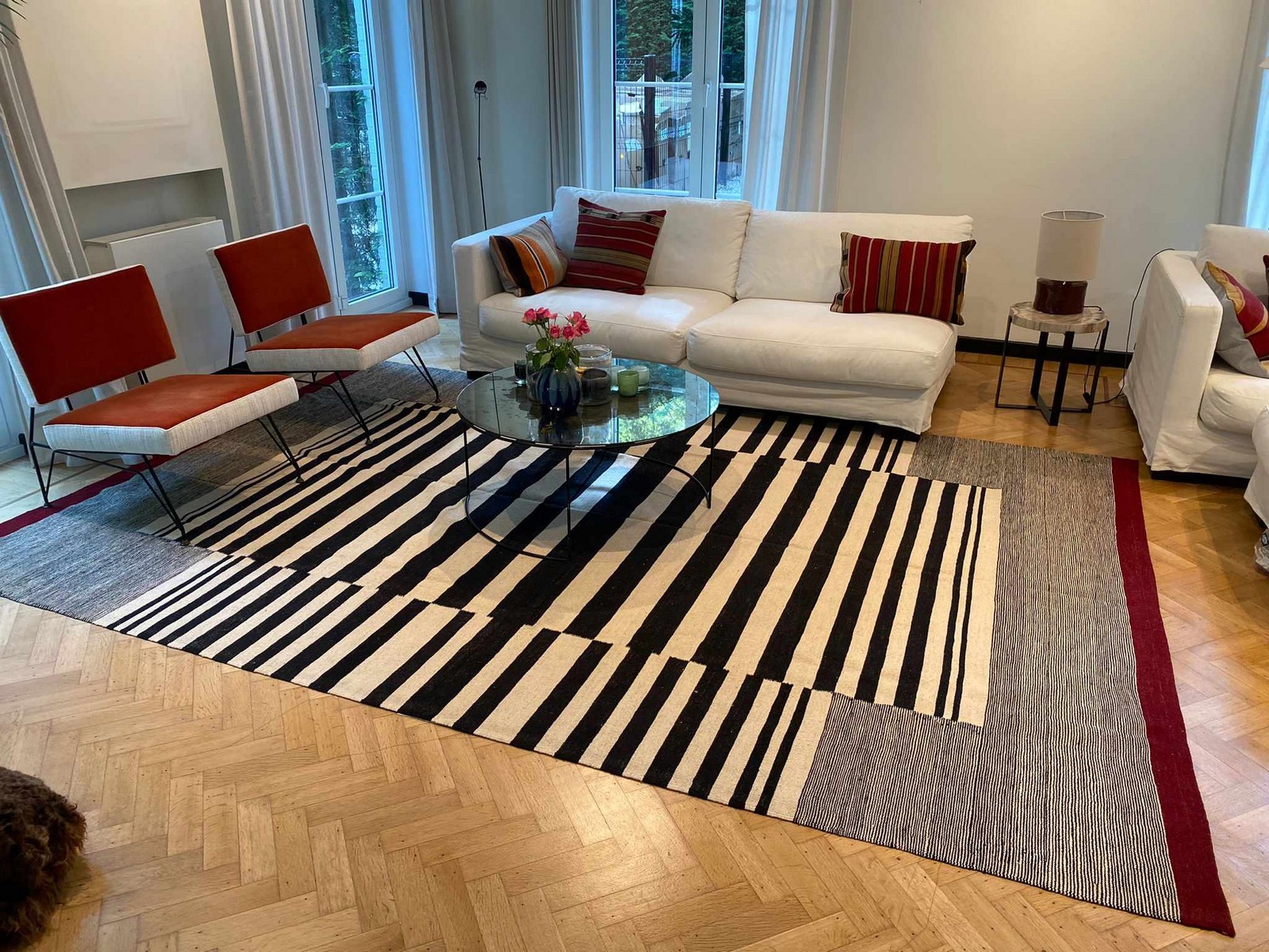 Woonkamer met witte bank, rode kussens, ronde glazen salontafel, gestreepte vloerkleed en grote ramen.